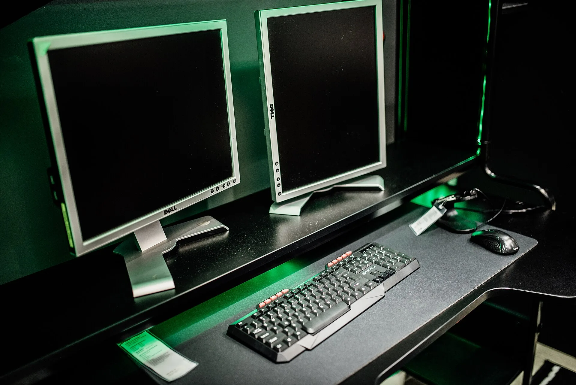 Two monitors sit on a desk in a gaming room. A computer keyboard and mouse are arranged neatly in front. The lighting is dim with green accents creating a tech environment.